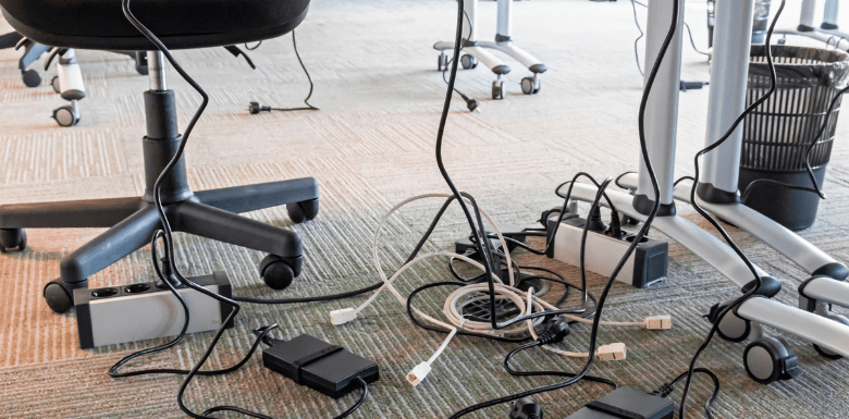 A picture of disorganized wires below a work desk on a beige carpeted floor.